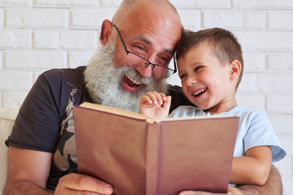 Bearded aged man and his grandson are having fun reading a book together while sitting in cozy armchair at home